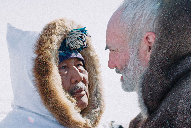 Indigenous man in parka face-to-face with a white man with white hair and beard.