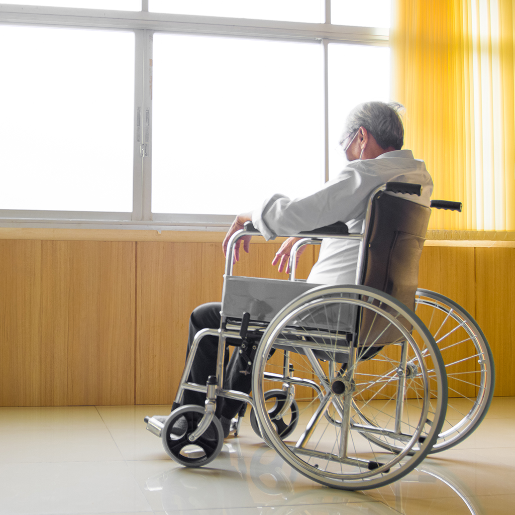 Elderly Asian person is sitting on a wheelchair, alone by a window.