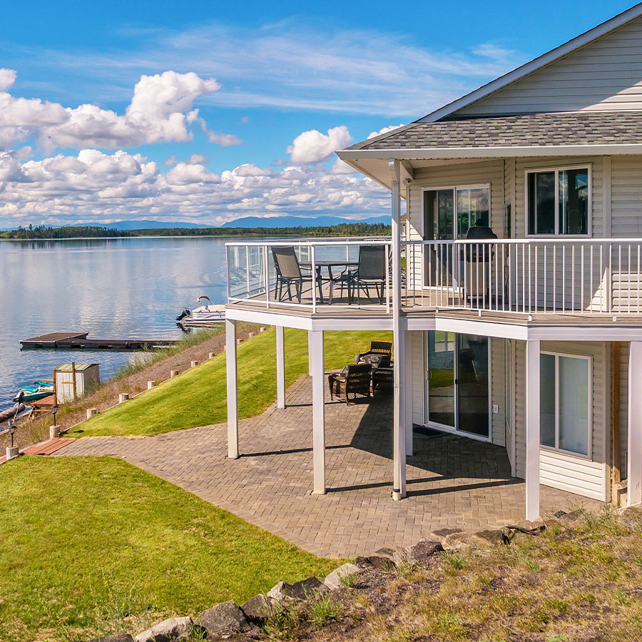 A large two-story house with beige vinyl siding, a shingled roof, a large outdoor deck and patio, with outdoor furniture situated on a freshwater lake. There is a a dock with a sitting area and equipment for water sports.