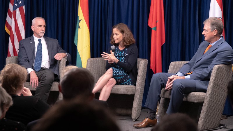 Three people sit in chairs with flags behind them