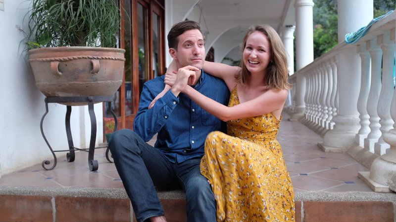 Two Peace Corps Volunteers sit on tiled steps. They are leaning on each other and smiling.