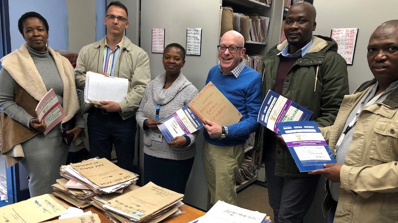 White male volunteer stands in health clinic with South African colleagues with files and all are smiling.
