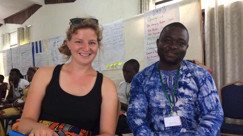 A woman and a man sit next to one another, both wearing colorful prints. Visible in the background are flip charts with writi