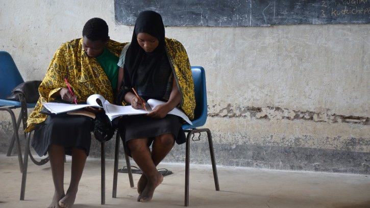 Two Malawian sit together and write in notebooks in a classroom.