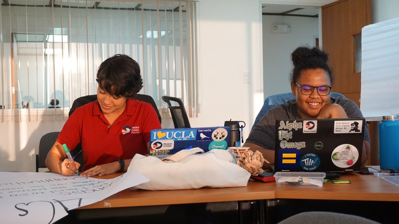 Diane smiles as she works on her computer. To the left a coworker works on a poster.