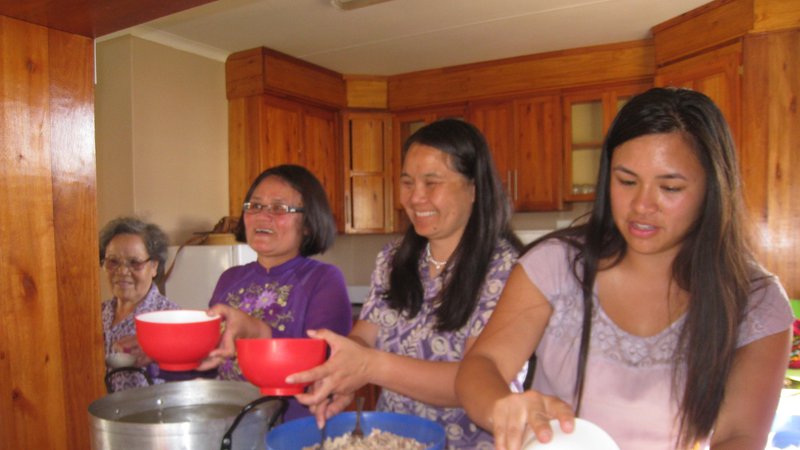 Four Asian American women stand side by side in a kitchen. They are cooking together.