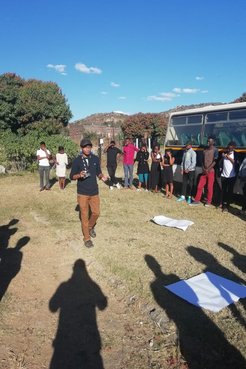 An male African American Peace Corps volunteer stands outside on the grass surrounded by South African teenagers next to a bu