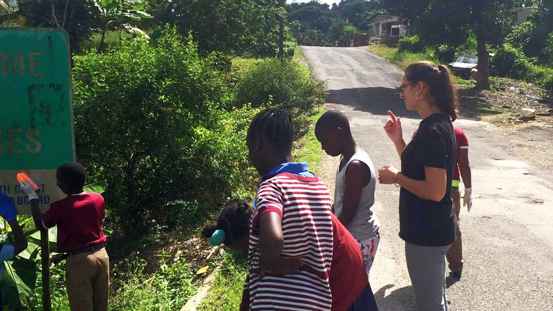 A white school teacher standing with two primary school students in brown watch other students clean a sign