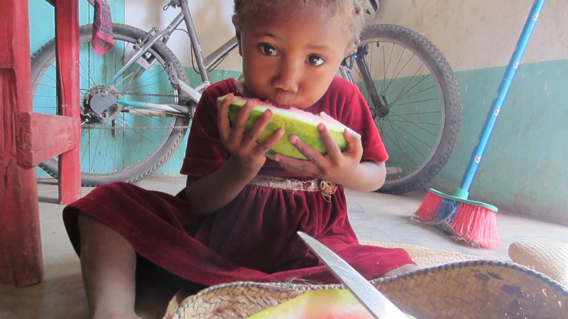 A young girl in Madagascar sits on the floor eating a large chunk of watermelon.