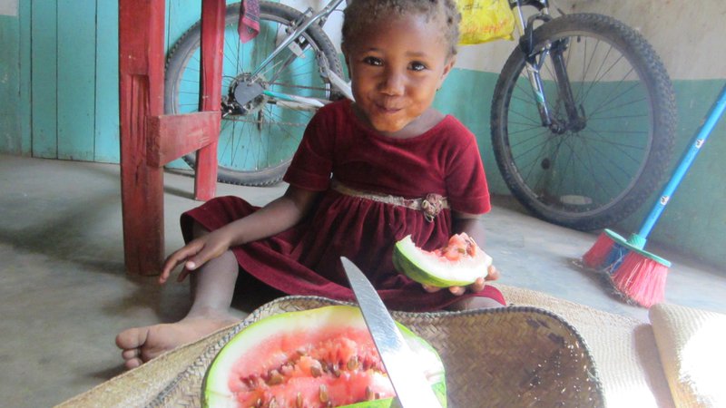A young girl in Madagascar sits on the floor, smiling. There is a large piece of watermelon in her hand.