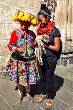 A Peace Corps Volunteer stands next to a woman in traditional Peruvian dress. The Volunteer is holding a baby sheep in her arms and smiling.