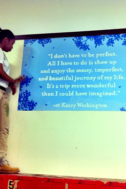 Brittany stands to the left of the frame on top of a cabinet. She is beside a freshly painted mural with a quote from Kerry Washington that reads, "I don't have to be perfect. All I have to do is show up and enjoy the messy, imperfect and beautiful journey of my life. It's a trip more wonderful than I could have imagined."