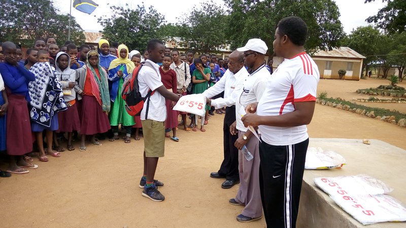Four adult people stand infront of a crowd of Tanzanian school children holding malaria net materials.