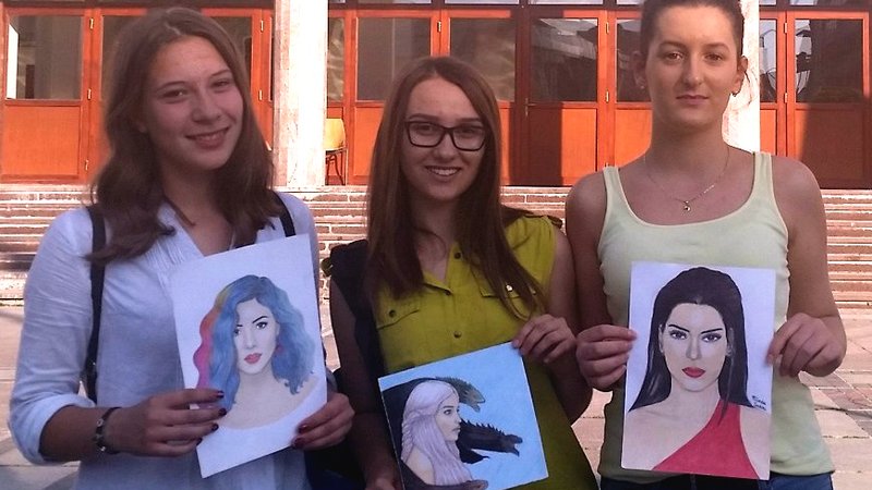 Three young women holding self-portrait paintings.