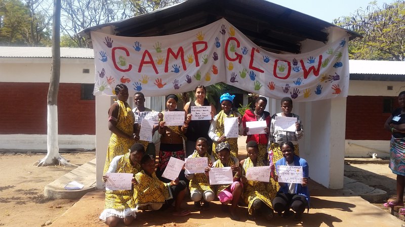 A Peace Corps Volunteer stands proudly with her Camp Glow participants under a banner.