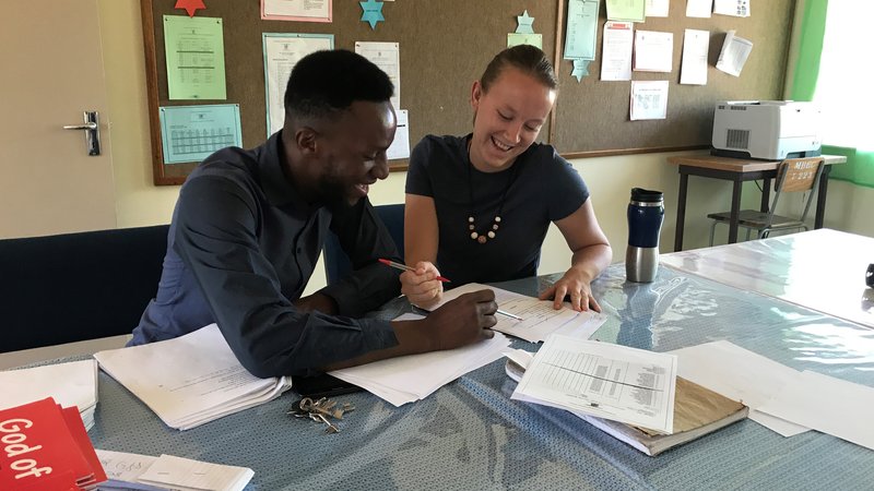 A man and a woman sit seated at a table. The table is strewn with papers, and they look to be working on an assignment togeth