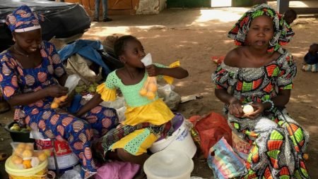 Two Gambian women sit outside at a market selling oranges.