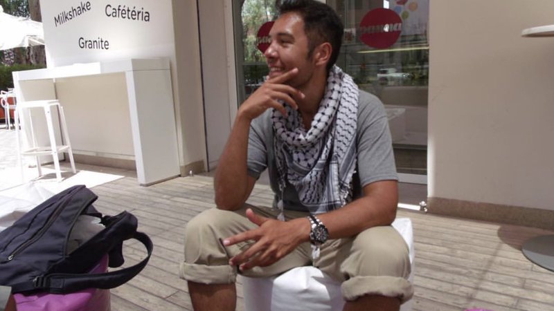 A man sits outside of a white ice cream shop. He is looking off camera and smiling.
