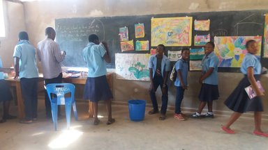 Students in blue uniforms stand at the front of the room looking at their paintings displayed on the black board