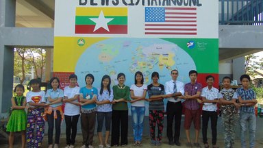 Teachers and students stand and link arms in front of a large, painted world map.