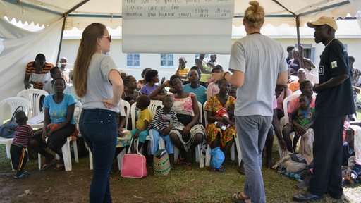 A male and female Volunteer stand with a male Ugandan counterpart and deliver a lesson about malaria to a group of about twenty people under the shade of a tent.