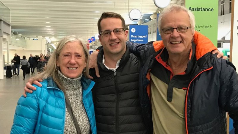 A young white male stands between his mother and father outside. All three smile.