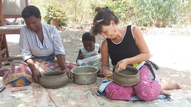 Birgit sits on a bamboo mat with a Malawian woman and a child as they hold and mold clay pots in their laps