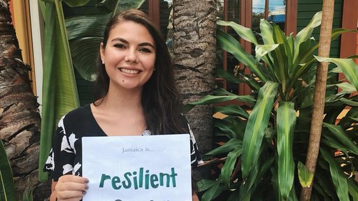 Turkish Female Peace Corps Volunteer holds a sign that reads "Jamaica is Resilient" in front of an orange building.