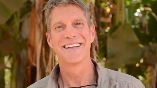 An older male Volunteer smiles at the camera while standing under a banana tree.