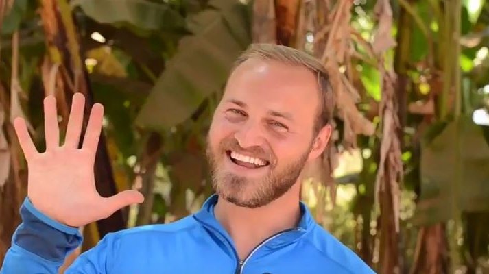 A blonde male Volunteer holds up his hand in greeting with a smile.